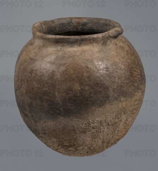 Kitchen pot, unknown northern Mesopotamian workshop. Early Jazira III period ca. 2600–ca. 2350 BC. A kitchen pot with an egg-shaped belly and rounded bottom, terminating in a low, wide cylindrical neck with a rounded, slightly tilted spout. Two thickened, horizontal crescent-shaped handles are attached on opposite edges. Outer walls are intentionally roughened at the bottom up to half the vessel's height and polished above. Traces of soot are visible at the bottom and slightly above. The clay contains a rich admixture of crushed quartz. Tell Rad Shakra northeastern Syria; Northern Mesopotamia. Vessel; height 23.3 cm, width 23.3 cm., A rounded earthenware vessel with a wide mouth and thick rim sits against a neutral gray background, showing a weathered surface in shades of brown, tan, and dark gray with areas of lighter beige and subtle mottling; the body displays rough, textured patches and smoother worn sections, with visible irregularities, faint cracks, and a somewhat darker banding...