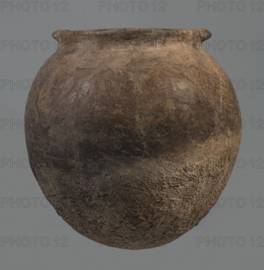 Kitchen pot, unknown northern Mesopotamian workshop. Early Jazira III period ca. 2600–ca. 2350 BC. A kitchen pot with an egg-shaped belly and rounded bottom, terminating in a low, wide cylindrical neck with a rounded, slightly tilted spout, to which two thickened, horizontal crescent-shaped handles are attached on opposite edges. Outer walls intentionally roughened at the bottom up to half the height and polished above. Traces of soot visible at the bottom and slightly above. Fabric contains a rich admixture of crushed quartz. Syria; Tell Rad Shakra northeastern Syria. Northern Mesopotamia place of manufacture. Vessel; height 23.3 cm, width 23.3 cm., A rounded, weathered clay vessel with a narrow, slightly flared rim dominates the image, showing a rough, pitted surface texture and irregular patches of wear; the vessel exhibits a palette of brown tones ranging from light tan to dark brown with areas of gray and muted ochre, and subtle variations in shading that emphasize its uneven, age...