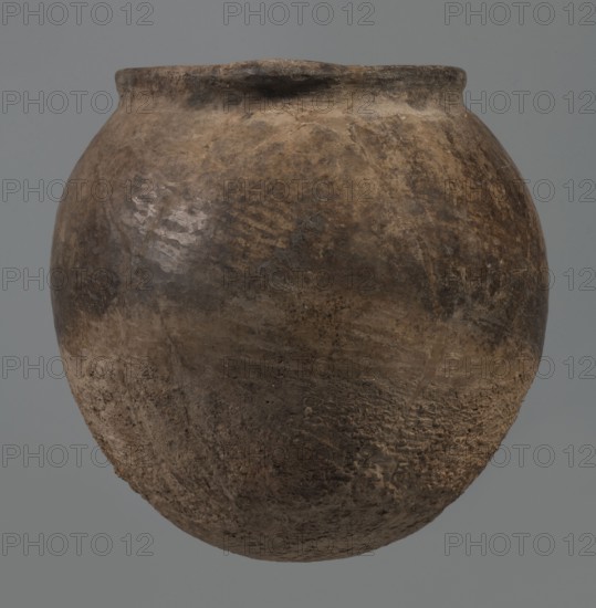 Kitchen pot, unknown northern Mesopotamian workshop; Early Jazira III period ca. 2600–ca. 2350 BC. Egg-shaped belly with rounded bottom, terminating in a low, wide cylindrical neck with a rounded, slightly tilted spout; two thickened, horizontal crescent-shaped handles attached on opposite edges. Outer walls intentionally roughened at the bottom up to half the vessel height and polished above. Traces of soot visible at the bottom and slightly above. Fabric contains a rich admixture of crushed quartz. From Tell Rad Shakra northeastern Syria, Northern Mesopotamia. Vessel; height 23.3 cm, width 23.3 cm., A rounded earthen vessel with a wide body and slightly narrowed rim, showing a rough, textured surface with patches of lighter and darker areas; colors visible include brown, tan, beige, gray, and hints of black, and the vessel displays irregular mottling, worn spots, and a few areas with apparent scraping or pitting against a plain gray background.