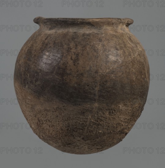 Kitchen pot, unknown northern Mesopotamian workshop. Early Jazira III period ca. 2600–ca. 2350 BC. A kitchen pot with an egg-shaped belly and a rounded bottom, terminating in a low, wide cylindrical neck with a rounded, slightly tilted spout, to which two thickened, horizontal crescent-shaped handles are attached on opposite edges. The outer walls are intentionally roughened at the bottom up to half the height of the vessel and polished above. Traces of soot are visible at the bottom and slightly above. The fabric contains a rich admixture of crushed quartz. Syria; Tell Rad Shakra northeastern Syria. Northern Mesopotamia place of manufacture. Vessel; height 23.3 cm, width 23.3 cm., A rounded clay vessel with a wide belly and a slightly flared rim, shown against a plain gray background, displaying an overall weathered surface with a mix of brown, dark brown, and black tones; the upper area has smoother darker patches and some lighter worn spots, while the lower area appears rougher and ...