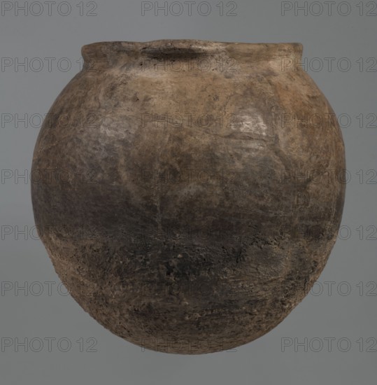 Kitchen pot, unknown northern Mesopotamian workshop. Kitchen vessels, clay vessel, crescent-shaped handles. Early Jazira III period ca. 2600–ca. 2350 BC. A kitchen pot with an egg-shaped belly and a rounded bottom, terminating in a low, wide cylindrical neck with a rounded, slightly tilted spout, to which two thickened, horizontal crescent-shaped handles are attached on opposite edges. The outer walls are intentionally roughened at the bottom up to half the height of the vessel and polished above. Traces of soot are visible at the bottom and slightly above. The clay contains a rich admixture of crushed quartz. Syria; Northern Mesopotamia; Tell Rad Shakra northeastern Syria. Vessel; height 23.3 cm, width 23.3 cm., A large rounded clay vessel with a bulbous shape and a slightly flared rim, shown against a plain neutral background; the surface of the vessel appears weathered and uneven with a rough texture and patches of wear, displaying a mix of earthy tones including brown, tan, gray, a...