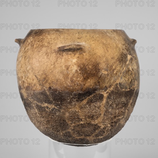 Kitchen pot, unknown northern Mesopotamian workshop; kitchen vessels. A kitchen pot with an egg-shaped belly with a rounded bottom, slightly inward-sloping walls, terminating at the top in a rim diagonally cut inward. A few centimeters below the rim, four symmetrically placed horizontal crescent-shaped handles are attached. The outer walls are intentionally roughened at the bottom up to 2/3 of the vessel's height, while polished above. The roughened part of the vessel is heavily sooted. The fabric contains a rich admixture of crushed quartz. Clay vessel; 1st half of the 3rd millennium BC, Early Jazira III period. From Tell Rad Shakra northeastern Syria, Northern Mesopotamia. Vessel; height 23.5 cm, width 25.3 cm, weight 2.6 kg., A rounded, weathered pot with a bulbous shape sits against a plain light gray background, showing a textured surface with a mottled palette of tan, brown, dark brown, and beige; the pot has a few small protrusions near its rim, an uneven rim edge, and visible a...