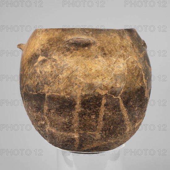 Kitchen pot, unknown northern Mesopotamian workshop; kitchen vessels. A kitchen pot with an egg-shaped belly with a rounded bottom, slightly inward-sloping walls, terminating at the top in a diagonally cut inward rim. A few centimeters below the rim four symmetrically placed horizontal crescent-shaped handles are attached. The outer walls are intentionally roughened at the bottom up to 2/3 of the vessel's height and polished above; the roughened part is heavily sooted. The fabric contains a rich admixture of crushed quartz. 1st half of the 3rd millennium BC; Early Jazira III period. Findspot: Tell Rad Shakra northeastern Syria. Northern Mesopotamia place of manufacture. Vessel. Height 23.5 cm; width 25.3 cm; weight 2.6 kg., A rounded, weathered pot sits against a plain light gray background, showing a textured surface with a mottled pattern of tan, brown, and darker brown areas; the pot has an uneven rim and a small protrusion on one side, with visible cracks and repaired seams across ...