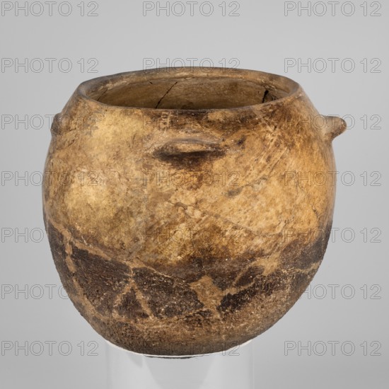 Kitchen pot, unknown northern Mesopotamian workshop; kitchen vessels. A kitchen pot with an egg-shaped belly with a rounded bottom, slightly inward-sloping walls, terminating at the top in a rim, diagonally cut inward. A few centimeters below the rim, four symmetrically placed horizontal crescent-shaped handles are attached. The outer walls are intentionally roughened at the bottom up to 2/3 of the vessel's height, while polished above. The roughened part of the vessel is heavily sooted. The fabric contains a rich admixture of crushed quartz. 1st half of the 3rd millennium BC; Early Jazira III period. Tell Rad Shakra northeastern Syria. Northern Mesopotamia place of manufacture. Vessel; height 23.5 cm, width 25.3 cm, weight 2.6 kg., A rounded, earthen vessel with a wide opening and a thick rim sits against a pale gray background; the pot displays a textured surface with a mottled palette of brown, tan, beige, and darker brown patches, showing areas of wear and irregular discoloration, ...