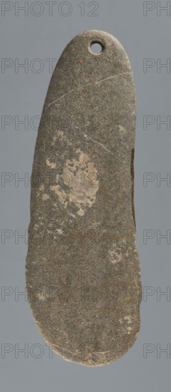 Whetstone, unknown northern Mesopotamian workshop; Early Jazira III / Early Dynastic III period ca. 2600–ca. 2350 BC; elongated, flat boulder with a biconically drilled hole at the top of the narrower end; tool whetstone, abrasive tool; material: stone; origin: Northern Mesopotamia Khabur basin, Tell Rad Shakra, northeastern Syria; dimensions: height 10.5 cm, width 3.5 cm, depth 0.7 cm., A vertically oriented elongated stone object with a rounded top and a slightly irregular bottom edge, featuring a circular hole near the top; the surface shows a coarse, mottled texture with patches of lighter beige areas and thin lighter veins against an overall gray and brown coloration, and the background is a flat muted gray.