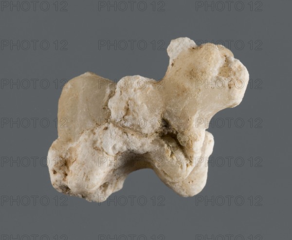 Pendant, unknown Northern Mesopotamian workshop, Early Jazira III period ca. 2600–ca. 2350 BC Early Dynastic III period. Figurine of an animal standing on all fours sheep?, separated straight dangling tail and large floppy ears on the head. A vertical hole drilled through the center of the back. The whole appearance is disturbed by white plastic mineral efflorescence. Origin: Northern Mesopotamia Khabur basin, Tell Rad Shakra northeastern Syria. Pendant; height 1.8 cm, width 2.3 cm, depth 1 cm., A close-up of an irregularly shaped rock or mineral specimen with a rough, bumpy surface and a mix of textures; the object shows a range of off-white, beige, and light tan tones with some slightly darker beige-brown patches, and appears porous in places against a flat, medium gray background.