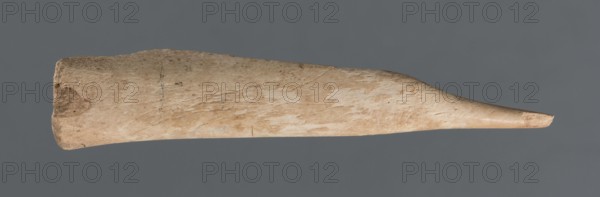 Awl;, Early Jazira III period ca. 2600–ca. 2350 BC / Early Dynastic III period ca. 2600–ca. 2350 BC; bone tool used as a tool; elongated awl, triangularly sharpened at one end; material: bone; function: awl; provenance/origin: Northern Mesopotamia, Al-Jazira Khabur basin, Tell Rad Shakra northeastern Syria; excavated; dimensions: height 8.5 cm, width 1.5 cm., A long tapered object with a rough textured surface lies horizontally against a gray background, showing beige and light brown tones with some slightly darker brown patches and subtle linear scratches; one end is broader and irregular with a darker brown spot while the opposite end narrows to a smoother, pointed tip with lighter beige coloring, and the overall surface displays variations of cream, tan, and muted brown.