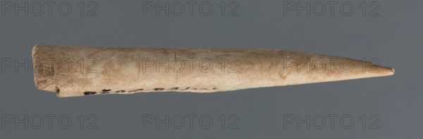 Awl, Early Jazira III / Early Dynastic III period ca. 2600–ca. 2350 BC; elongated awl, triangularly sharpened at one end; material: bone tool; function: awl; provenance/origin: Northern Mesopotamia, Al-Jazira Khabur basin, Tell Rad Shakra northeastern Syria; dimensions: height 8.5 cm, width 1.5 cm., A slender, tapered cylindrical object with a rounded blunt end and a pointed tip lies horizontally against a solid gray background; the object's surface is primarily beige with areas of light tan and subtle brown discoloration, showing a weathered, slightly rough texture with small scratches and shallow grooves, and a few darker brown spots near the blunt end.