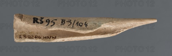 Awl, Early Jazira III / Early Dynastic III period ca. 2600–ca. 2350 BC. Elongated bone awl, triangularly sharpened at one end. From Tell Rad Shakra, Khabur basin, Northern Mesopotamia Al-Jazira, northeastern Syria. Function: tool. Dimensions: height 8.5 cm; width 1.5 cm., A narrow, elongated object with a pointed tapered end and a thicker blunt end, shown against a solid gray background; the object has an uneven, textured surface with visible chips and grooves, and displays a range of light beige, tan, and off-white colors with small areas of darker brown and faint greenish discoloration.