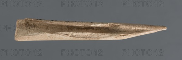 Awl, Early Jazira III / Early Dynastic III period ca. 2600–ca. 2350 BC. Elongated awl, triangularly sharpened at one end; material: bone; function: tool. Origins/place: Al-Jazira Northern Mesopotamia, Khabur basin, Tell Rad Shakra northeastern Syria. Dimensions: height 8.5 cm, width 1.5 cm., A single elongated, pointed object lies horizontally against a solid gray background, showing a few rough, textured surfaces with irregular pitting and scratches. The object displays a range of beige to light tan tones with some areas of darker brown discoloration and subtle off-white highlights. Its shape tapers to a sharp point at one end and has a slightly flattened, hollowed, or scooped channel running along its length, with uneven, chipped edges and granular surface detail visible along the sides.