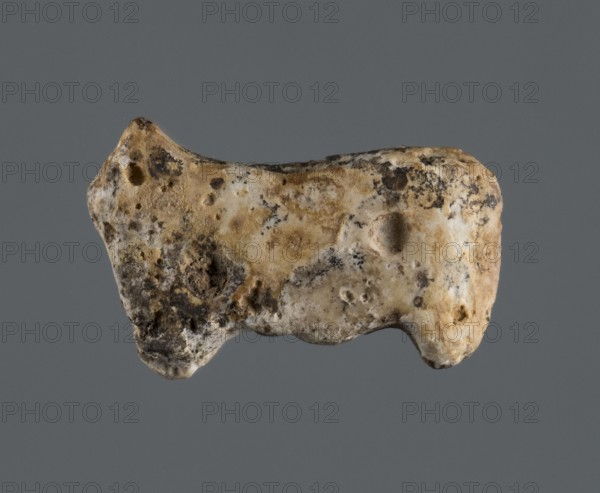 Pendant, unknown Northern Mesopotamian workshop, Early Jazira III period ca. 2600–ca. 2350 BC. Pendant or pin finial in the shape of a standing quadruped sheep?, head missing, plastically separated, straight hanging tail. On both sides of the animal, halfway up, there are two circular recesses drilled. A vertical hole is drilled through the centre of the torso. Excavated from Tell Rad Shakra, Khabur basin, northeastern Syria Northern Mesopotamia. Function: jewellery; associated with children's graves. Materials/decoration noted; height 1.8 cm, width 2 cm, depth 0.6 cm., A roughly irregular-shaped object with a pitted, porous surface suspended against a solid gray background; the object shows a mix of beige, tan, off-white, brown, and areas of dark gray or black mottling, with several small cavities and rough textured patches across its surface and rounded, uneven edges.
