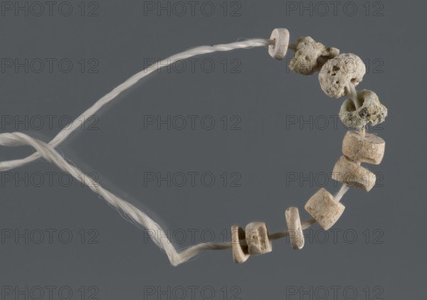 Fragment of a bracelet, unknown Northern Mesopotamian workshop, Early Jazira III period ca. 2600–ca. 2350 BC / Early Dynastic III period, beads, bracelets, disc-shaped beads, cylindrical beads, jewellery, children's graves. Ten small beads from disc-shaped to cylindrical of various diameters and heights. Tell Rad Shakra northeastern Syria, Northern Mesopotamia Khabur basin, Al-Jazira Syria/Iraq, ca. 2600–ca. 2350 BC. Decoration., A loop of twisted light-colored cord holds a group of small bead-like objects threaded onto it; several of the beads are smooth and rounded with a beige to light tan color and visible grain, while a few beads have rough, porous surfaces in pale off-white and light gray tones with some darker speckles. The cord appears creamy white and slightly glossy, and the background is a solid medium gray.