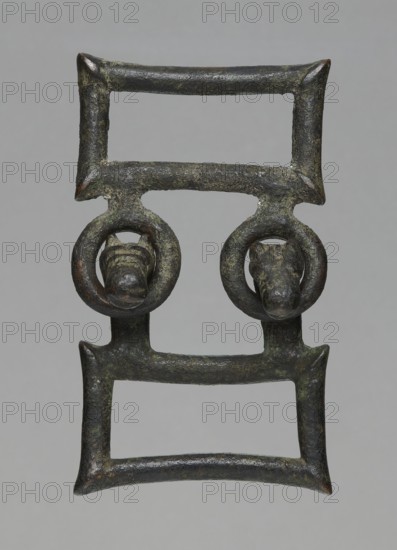 Buckle, bronze; two-part rectangular form with joints; one part with hooks shaped as zoomorphic animal heads; decoration; 7th century BC, Etruria; length 13 cm total, height 7.5 cm, thickness 4 cm, weight 0.374 kg, length 7.2 cm single element., An aged metal object with a rectangular, ladder-like form composed of two parallel bars at top and bottom connected by vertical segments, featuring a pair of circular loops suspended from the middle bar each encircling a small sculpted animal head; the surface shows a weathered, rough texture with patches of dark green and brown patina, along with areas of darker gray and hints of reddish-brown.
