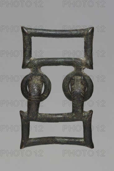 Buckle, Bronze buckle consisting of two rectangular parts with joints; one part has hooks shaped as zoomorphic decoration animal heads. 7th century BC, Etruria. Decoration. Total length 13 cm, height 7.5 cm, thickness 4 cm, weight 0.374 kg; length of single element 7.2 cm., An aged metal object with a rectangular double-frame structure connected by horizontal bars, showing a mottled patina in shades of dark green, brown, and black; two ring-shaped elements hang from the upper crossbar, each supporting a stylized animal head with elongated snouts and carved ridges, the heads and rings sharing the same greenish-brown metallic surface with patches of darker corrosion and lighter verdigris, all set against a plain neutral gray background.