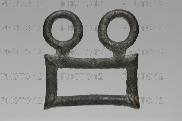 Buckle, Bronze buckle consisting of two parts in a shape similar to rectangles with joints. One part of the buckle has hooks in the form of zoomorphic decoration animal heads. 7th century BC, Etruria. Decoration. Total length 13 cm, height 7.5 cm, thickness 4 cm, weight 0.374 kg; length 7.2 cm single element., An object with a rough, weathered surface in dark green and brown tones is shown against a neutral gray background; it has a rectangular lower frame with slightly flared corners and two circular looped forms attached to the top corners, the material appearing corroded and pitted with patches of lighter green and hints of reddish-brown.