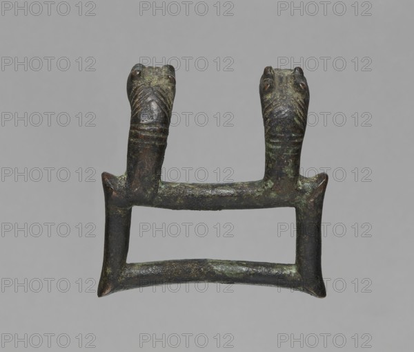 Buckle, Bronze buckle consisting of two parts in a shape similar to rectangles with joints. One part of the buckle has hooks in the form of zoomorphic decoration animal heads. 7th century BC, Etruria. Decoration. Length 13 cm total; height 7.5 cm; thickness 4 cm; weight 0.374 kg; length 7.2 cm single element., A small bronze object shaped like a rectangular frame with a horizontal bar across the middle and two vertical supports rising from the top of the frame, each support ending in a stylized animal head with carved patterns on the snouts; the surface shows a mottled patina with shades of green, dark brown, and hints of black, and the background is a uniform light gray.