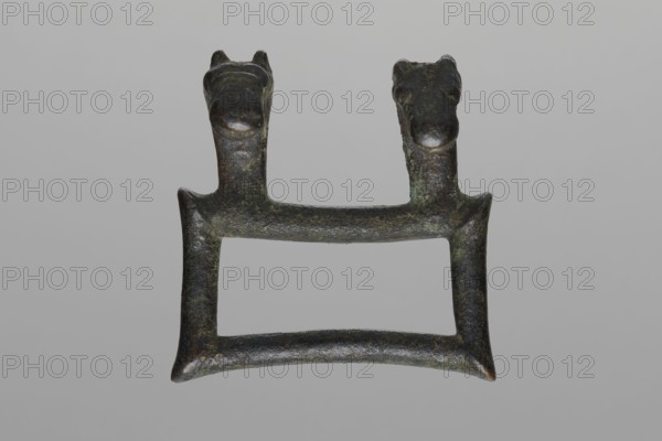 Buckle, Bronze buckle consisting of two parts in a shape similar to rectangles with joints. One part of the buckle has hooks in the form of zoomorphic decoration animal heads. 7th century BC, Etruria. Decoration. Length 13 cm total, height 7.5 cm, thickness 4 cm, weight 0.374 kg, length 7.2 cm single element., An aged metal object with a rectangular open center and a slightly curved lower bar, showing a rough, pitted surface in dark green, dark brown, black, and hints of lighter green patina, topped by a pair of short vertical posts each ending in a rounded animal-like head with small ears and snouts, all set against a plain light gray background.