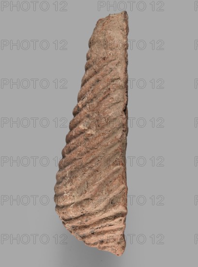 Votive offering in the form of a womb, votive offerings, body parts, Ex-voto in the form of a womb, 332–30 BC Hellenistic period, Caere place of origin; votive offering; height 4.5 cm, length 13.2 cm, width 9 cm, weight 0.27 kg., A roughly triangular, vertically oriented fragment with a textured, ridged surface and irregular edges; the surface shows a repeating diagonal groove pattern creating a spiral-like effect, with a rough, grainy texture and small pits; visible colors include shades of reddish-brown, tan, beige, gray, and small darker specks; the background is a flat medium gray.