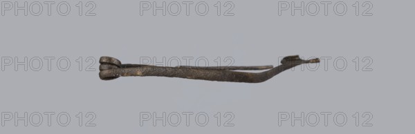 Fibula, bronze Etruscan fibula with a bent ring made of flatly beaten wire, passing in a three-turn spiral into a thin pin. Small sheath made of a sheet of metal with a rolled-up lower edge. Condition: green patina, hook plate partially broken off, needle bent, without tip. 6th–5th century BC, Southern Etruria. Height 6.7 cm., A single elongated metal object is centered against a plain light gray background, showing a dark brown and black surface with patches of lighter brown and slight metallic sheen; one end has a twisted loop-like cluster with rounded protrusions, while the opposite end tapers into a flattened curved tip; the rod between the ends appears irregular and textured with rough, mottled coloring and subtle variations of dark gray and rusty brown.