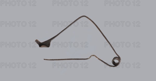 Fibula, Bronze Etruscan fibula with a bent ring made of flatly beaten wire, passing in a three-turn spiral into a thin pin. Small sheath made of a sheet of metal with a rolled-up lower edge. Condition: green patina, hook plate partially broken off, needle bent, without tip. 6th–5th century BC, Southern Etruria. Height 6.7 cm., A thin, irregularly shaped metal object with a dark brown to black patina is shown against a pale gray background; the object has a long, tapered arm that curves upward into an angled peak, a coiled loop at one end, a flattened thicker section near the opposite end, and a slender straight extension with a slight bend, displaying rough, textured surfaces and subtle variations of brown, black, and metallic gray.