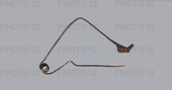 Bronze Etruscan fibula with a bent ring made of flatly beaten wire, passing in a three-turn spiral into a thin pin. Small sheath made of a sheet of metal with a rolled-up lower edge. Condition: green patina, hook plate partially broken off, needle bent, without tip. 6th–5th century BC, Southern Etruria. Height 6.7 cm., A thin, irregularly shaped metallic wire object with a coiled loop at one end and a slightly flattened, thicker section at the opposite end, shown against a pale gray background; the metal appears dark brown to black with areas of rusty orange and lighter brown highlights along its worn surface, and the object forms a triangular outline with a gently arched top segment and a lower straight segment that has a small angular bend.