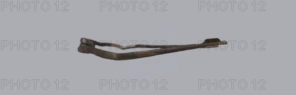 Fibula, Bronze Etruscan fibula with a bent ring made of flatly beaten wire, passing in a three-turn spiral into a thin pin. Small sheath made of a sheet of metal with a rolled-up lower edge. Condition: green patina, hook plate partially broken off, needle bent, without tip. 6th–5th century BC, Southern Etruria. Height 6.7 cm., A single rusted metal object shaped like a thin, irregular loop is centered against a smooth light gray background; the object appears textured and worn with dark brown and black tones and subtle areas of lighter brown and gray, showing uneven edges and a slightly flattened section at one end.