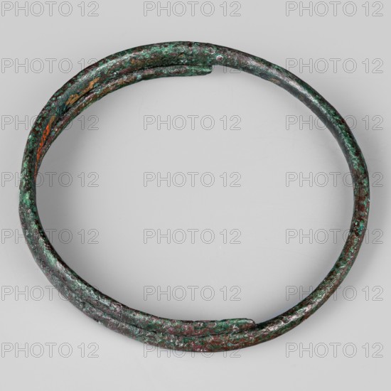 Bracelet. Made of flattened wire with thinning, overlapping ends. 9th–8th century BC; Villanovian period, Etruria. Body ornament; weight 46 g; height 1.2 cm; diameter 8.5 cm., A circular metal band with a rough, pitted surface lies against a light gray background; the band shows a mottled mix of green and brown patina with patches of darker brown and hints of reddish tones, and a few small areas where the metal appears worn or brighter.