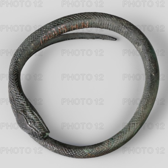 Bracelet. Made of flattened wire with thinning, overlapping ends. 9th–8th century BC; Villanovian period, Etruria. Body ornament; weight 46 g, height 1.2 cm, diameter 8.5 cm., A coiled bracelet or ring shaped like a snake with a textured, scale-like surface visible across most of its body, showing wear and small surface pitting; the snake head is detailed with carved features and a small rounded eye, and the tail tapers to a thinner end that lies near the inner curve. The object displays a mix of dark metallic tones with areas of greenish patina and spots of brownish corrosion, set against a light gray background.
