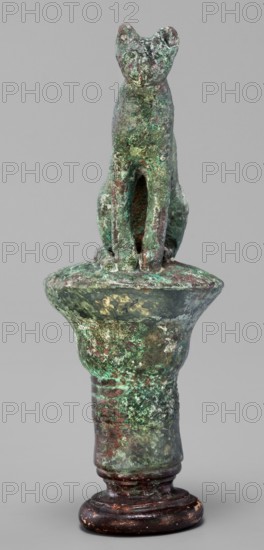 Cat sitting on a papyrus column;, Egyptian art, Late Period, 664–332 BC. Bronze figurine. Height 11 cm; width 4.5 cm; weight 0.314 kg. Subjects: cats; capitals., A small sculptural object depicting a seated animal figure atop a pedestal, with a textured surface showing a patina of green and turquoise alongside areas of brown, dark bronze, and hints of reddish tones; the animal has a simplified head with upright ears and a narrow body with visible front limbs, and the pedestal beneath it flares outward near the top and tapers to a rounded base that is darker brown with worn lighter spots.