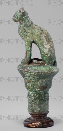 Cat sitting on a papyrus column, cats, Scen.Gal.Eg., capitals, Egyptian art, 664–332 BC; Late Period, Egypt, figurine, height 11 cm, width 4.5 cm, weight 0.314 kg, A weathered sculptural object showing a seated animal figure atop a rounded platform on a turned base, with a rough, textured surface. The animal has a stylized head, ears, a curved back, and front legs that extend down to the platform, leaving an open space beneath its body. The surface displays a mottled mix of green and verdigris tones with patches of brown and dark reddish-brown, and areas of lighter off-white and pale grey encrustation. The platform and the upper portion of the pedestal share the same mottled green and brown patina, while the lower turned base is dark reddish-brown with worn areas revealing lighter beige and pale spots. The background is a flat, neutral grey.