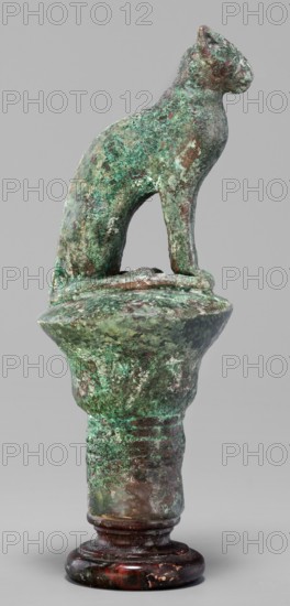 Cat sitting on a papyrus column; creator, cats; Scen.Gal.Eg.; capitals; Egyptian art; 664–332 BC Late Period, Egypt; figurine; height 11 cm; width 4.5 cm; weight 0.314 kg., A weathered sculptural object resembling an animal figure perched atop a rounded pedestal, with a mottled surface showing shades of green, turquoise, brown, and traces of white; the animal form has a curved back, upright head, and slender legs, and the pedestal beneath it widens into a flared section above a cylindrical shaft that rests on a polished base exhibiting darker reds, browns, and black tones.