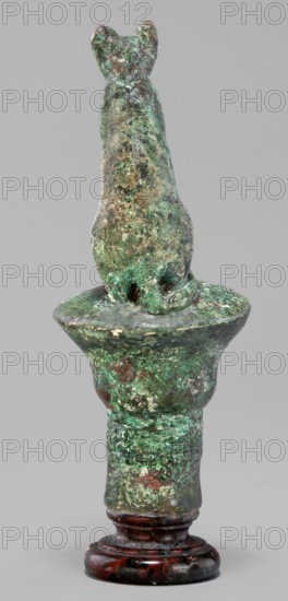 Cat sitting on a papyrus column, cats; Scen.Gal.Eg.; capitals; Egyptian art; 664–332 BC Late Period, Egypt; figurine; height 11 cm, width 4.5 cm, weight 0.314 kg., An upright sculptural object resembling an animal figure perched on a rounded pedestal, shown from behind against a plain gray background, with an overall weathered surface showing a mottled mix of green, brown, and turquoise patina, darker brown and reddish areas, and some lighter worn spots; the base appears glossy dark reddish-brown with black veining.