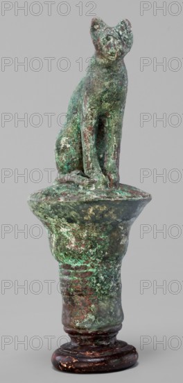 Cat sitting on a papyrus column; maker, cats; Scen.Gal.Eg.; capitals; Egyptian art; 664–332 BC, Late Period, Egypt; figurine; height 11 cm; width 4.5 cm; weight 0.314 kg, A weathered small sculpture of a seated cat figure atop a rounded pedestal, showing a rough textured surface with visible corrosion and patina in shades of green and turquoise mixed with areas of brown and reddish-brown, mounted on a darker rounded base that is glossy and deep brown with small worn spots.