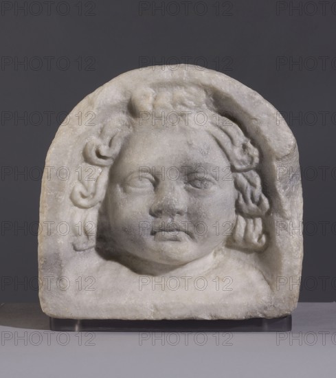 Fragment of a sarcophagus lid, unknown Italian workshop. Fragment of a sarcophagus lid depicting the face of a child with a round face and prominent cheeks. The depiction is frontal, with almond-shaped eyes, disproportionately large compared to the rest of the face, with carefully sculpted eyelids and pupils. The nose is also large, triangular. The hair above the forehead is gathered in a thick bun. Around the head, curls form harmoniously on both sides, covering the ears. The neck is short and stocky. Based on the figure's appearance, it is difficult to determine the child's gender, although the depiction is portrait in nature. This is a fragment of a rare type of sarcophagus lid, decorated with a frieze of semicircular parapets depicting various figurative scenes and themes, representations of the deceased. The portrait features and attention to physiognomic detail may suggest that this is a depiction of a deceased child; similar Cupid putti heads also decorate this type of sarcophag...