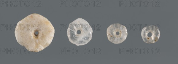 Set of four disc-shaped beads of different sizes, unknown northern Mesopotamian workshop; beads, disc-shaped, jewellery; likely from children's graves; ca. 2600–ca. 2350 BC, Early Jazira III period Early Dynastic III; Northern Mesopotamia Khabur basin, Tell Rad Shakra, Northeastern Syria; decoration., A row of four round, donut-shaped translucent objects arranged against a solid grey background, each with a central hole and irregular, slightly rough edges; the leftmost piece is larger and milky off-white with beige and pale tan speckles and a crystalline texture, the next is a clear to cloudy pale white with subtle internal fractures, the next is slightly smaller and translucent with faint beige inclusions and a glossy surface, and the rightmost is the smallest, mostly clear with small tan flecks and a shiny, irregular surface.