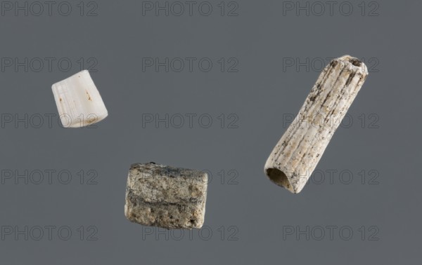 Set of three beads, unknown Northern Mesopotamian workshop; Early Jazira III period ca. 2600–ca. 2350 BC; beads, tubular beads, jewellery; Al-Jazira Syria; Iraq; Mesopotamia; used in children's graves; Set of three different tubular beads: one a naturally broken from a snail shell, two b, c cut from the shell of an unidentified snail. Bead b has a rounded triangular cross-section; c. 2600–c. 2350 BC; Northern Mesopotamia Khabur basin, Tell Rad Shakra northeastern Syria; decoration., A gray background with a few small cylindrical and tubular objects suspended against it; one object is white with faint brown spots and fine linear ridges, another is off-white with darker speckles and vertical grooves and a hollow end, and a third is a mottled gray-beige piece with a rough textured surface.