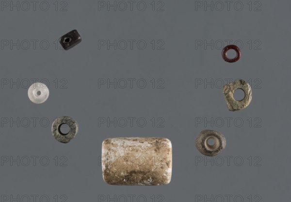 Bead set, Rad Shakra northeastern Syria, Polish excavations in northeastern Syria, mid-3rd millennium BC, Tell Rad Shakra, decoration, A group of small round and cylindrical objects arranged against a plain gray background, including a rectangular bead with a mottled tan and white surface near the bottom center, a few ring-shaped beads in colors such as dark brown, red, translucent white, muted green, and gray scattered around, and a darker cylindrical bead near the top left, all showing varied textures from smooth to rough.
