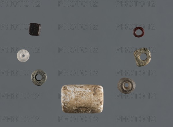 Bead set, Rad Shakra northeastern Syria, Polish excavations in northeastern Syria, mid-3rd millennium BC, Tell Rad Shakra, decoration, A plain gray background with a group of small objects arranged centrally, including a larger cylindrical bead with a worn beige and white surface near the bottom center, a few ring-shaped beads with rough textured surfaces in shades of dark greenish-gray, brown, and translucent white scattered to the left and right, a small dark square bead near the upper left, and a small dark red circular bead near the upper right.