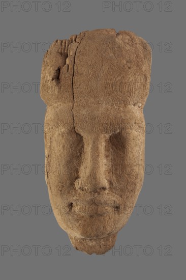 Sarcophagus mask with fragment of a wig above the forehead and a preserved piece of the neck. Very narrow face. No color. ca. 1069–332 BC; Third Intermediate Period–Late Period, Egypt. Sarcophagus mask., A carved face-shaped object in brown tones centered against a uniform gray background, showing textured, weathered surfaces and visible grain patterns; the features include a broad forehead, deep-set eye sockets, a pronounced nose, and a closed mouth with subtle lip contours, with cracks and rough edges along the top and sides and variations of lighter and darker browns highlighting the carved details.