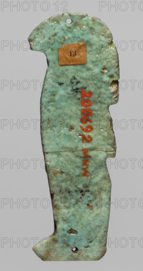 Amulet - Kebehsenuf element of a mummy net, Kebehsenuf myth., Sons of Horus myth., mummies, grave goods, Presumably an amulet from a mummy net, 664–332 BC; Late Period, Egypt, amulet, height 7.1 cm, width 2.6 cm, A vertical, elongated artifact shaped like a stylized human figure in profile, rendered in a bluish-green material with a mottled surface showing lighter and darker patches and areas of brownish discoloration; the figure has a rounded head with a small hole near the top, an indented area where a face in profile would be, and a widened lower section forming a foot-like projection with another small hole; a torn rectangular fragment of brown material is affixed near the upper section, and a vertical sequence of small orange shapes runs down the center of the object's front; the surface shows several small rough spots and pits, and the background behind the object is a plain, neutral light gray.