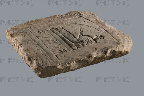 Rectangular tombstone depicting a young man in a long robe in a semi-reclining position on a bed with a mattress and two pillows. He holds a flat bowl or patera in his right hand. The bed legs are decorated; sacrificial vessels stand under the bed. A vine grows from the footrests on the right. The vine and the vessels are shallowly engraved, while the man is in high relief. Inscription in the lower part lists the deceased's name: Drakon, son of Apion. 2nd–4th century, Terenuthis. Bas-relief tombstone with Greek inscription. Dimensions: height 33 cm, width 30 cm, depth 5.5 cm., A roughly rectangular stone slab with a weathered, uneven surface and rough edges rests against a plain gray background; the slab is primarily beige and light brown with areas of darker brown shading, and it features a recessed central panel carved with a relief of a reclining human figure draped in flowing garments and surrounded by decorative incised lines and curved motifs; the carved area shows textured groov...
