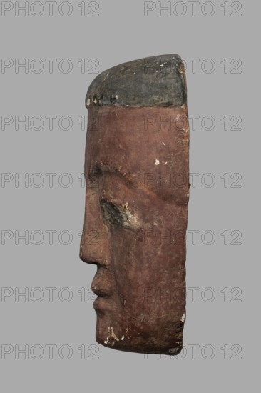 Outer sarcophagus mask, wig iconographic, forgeries, acquisition provenance, sarcophagus fragments, masks, faces. Sarcophagus mask with a flat fragment of a wig above the forehead. Elongated face, painted brown, high black eyebrow arches, black pupils filling almost the entire eyes, modeled mouth without marked lips. c. 747–332 BC; Late Period?, Egypt, forgery, A carved side profile of a human face rendered on a vertical rectangular block with a rounded top, showing a prominent nose, closed eye socket, and shaped lips; the surface appears textured and worn with chips and small areas of loss; the top portion is darker while the main face area is a reddish-brown with small patches of lighter wear and tiny spots of white; the background is a flat gray.