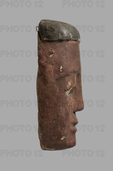 Sarcophagus mask with a flat fragment of a wig above the forehead. Elongated face, painted brown, high black eyebrow arches, black pupils filling almost the entire eyes, modeled mouth without marked lips. Outer sarcophagus mask; sarcophagus fragments; masks; faces. c. 747–332 BC Late Period?, Egypt. Forgery ; wig iconographic., A side view of a carved wooden mask or sculpture in front of a plain gray background showing a tall narrow face with a rounded top that is darker in color, a long brownish-red surface with worn and chipped areas revealing lighter patches, a pronounced nose and lips protruding in profile, subtle carved eye contours, a small metal loop near the top edge, and visible texture of aged wood with colors including brown, reddish-brown, dark gray, black, and small areas of beige.