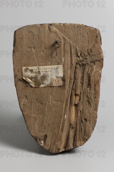 Outer sarcophagus mask, wig iconographic, forgeries, acquisition provenance, sarcophagus fragments, masks, faces. Sarcophagus mask with a flat fragment of a wig above the forehead. Elongated face, painted brown, high black eyebrow arches, black pupils filling almost the entire eyes, modeled mouth without marked lips. c. 747–332 BC; Late Period?, Egypt, forgery, A roughly shield-shaped wooden board with a weathered, textured surface showing grain, scratches, splits, and areas of rough, splintered wood; a small metal screw with a looped piece of thin wire is attached near the top; an aged rectangular paper label is affixed to the upper left area; the wood displays various brown tones from light tan to deeper brown and some darker grayish-brown streaks, while the background is a smooth light gray.