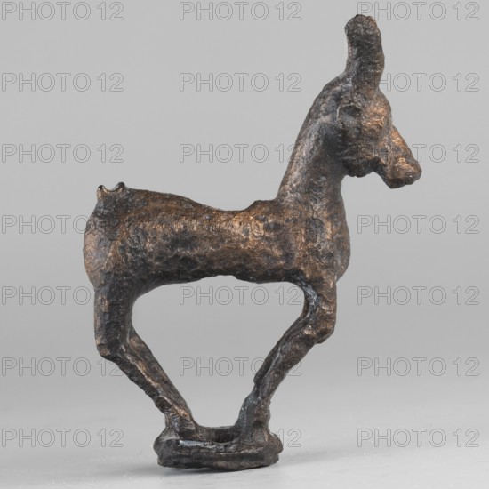 Emblem in the form of a figurine of a standing goat figurine of a standing gazelle with slightly bent legs on a ring-shaped base. Through the center of the gazelle's body runs a sleeve-shaped hole aligned with the hole in the ring, allowing the figurine to be mounted on, for example, a wooden handle. The animal's protruding ears contain holes that could have been used for hanging ornaments e.g., bells. Luristan bronze, unknown Luristan workshop; 1st half of the 1st millennium BC Iron Period II, Iran. Height 6.5 cm, width 4.8 cm, depth 1.7 cm., A small sculptural object shaped like an animal with a long neck and upright head, rendered in a dark metallic brown with areas of lighter bronze highlights, standing on a narrow base with two slender, curved legs forming an open space beneath the body; the surface is textured and irregular, showing rough, pitted details and subtle variations of brown and bronze tones against a neutral light gray background.