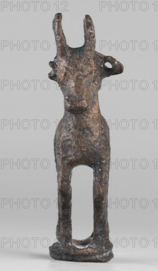 Emblem in the form of a figurine of a standing goat figurine of a standing gazelle with slightly bent legs on a ring-shaped base. Through the center of the animal's body runs a sleeve-shaped hole aligned with the hole in the ring, allowing the figurine to be mounted on, for example, a wooden handle. The animal's protruding ears contain holes that could have been used for hanging ornaments such as bells. Luristan bronze; unknown Luristan workshop. 1st half of the 1st millennium BC; Iron Period II, Iran. Height 6.5 cm, width 4.8 cm, depth 1.7 cm., A small sculpted figure of an animal standing upright on a rounded base, shown from the front, with two upright horn-like projections on its head and an ear-like protrusion on one side; the surface appears rough and irregular with visible texture and wear, and there are a few small fibrous particles caught on it. The figure's form narrows at the neck and widens at the chest, with slender front legs creating an open space between them above the ...