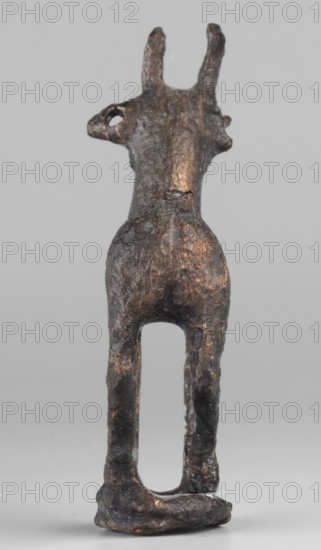Emblem in the form of a figurine of a standing goat or gazelle, unknown Luristan workshop. Figurine of a standing gazelle with slightly bent legs on a ring-shaped base. Through the center of the gazelle's body runs a sleeve-shaped hole, aligned with the hole in the ring; this arrangement of holes allowed the figurine to be mounted on, for example, a wooden handle. The animal's protruding ears contain holes that could have been used for hanging ornaments, e.g., bells. Luristan bronzes; animal figurine; 1st half of the 1st millennium BC, Iron Period II, Iran; place of manufacture: Luristan. Dimensions: height 6.5 cm, width 4.8 cm, depth 1.7 cm., A small sculptural object resembling the rear view of an animal with upright ears, made of a dark metallic material with patches of brownish-bronze highlights; the surface is rough and pitted with an uneven texture, showing a hollow area between elongated hind legs and a rounded rump above; the ears are narrow and upright, and there is a small ro...