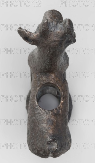 Emblem in the form of a figurine of a standing goat figurine of a standing gazelle with slightly bent legs on a ring-shaped base. Through the center of the animal's body runs a sleeve-shaped hole aligned with the hole in the ring, allowing the figurine to be mounted on, for example, a wooden handle. The animal's protruding ears contain holes that could have been used for hanging ornaments, e.g., bells. Luristan bronze, unknown Luristan workshop, 1st half of the 1st millennium BC Iron Period II, Luristan, Iran. Materials: bronze; dimensions: height 6.5 cm, width 4.8 cm, depth 1.7 cm., A dark brown and black metal object shaped like an animal form is shown from above against a light gray background, with a rounded head and two upward curving ear or horn-like protrusions, a textured surface with irregular dents and scratches, a large circular hole through the middle of its body, and a few areas of lighter brownish highlights and subtle greenish patina.