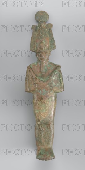 Figurine of Osiris depicted as a mummy-like standing figure wrapped in a shroud. The arms protruding from beneath the shroud are crossed over the chest in the so-called Upper Egyptian configuration. In his right hand, Osiris holds a scepter, while in his left he holds a whip. The head is adorned with an atef crown consisting of the White Crown and two ostrich feathers placed on the sides. The crown is topped with a solar disk and additionally adorned with a large uraeus with two lateral body coils and a long, wavy tail. The shroud fits softly to the body, highlighting the outline of the back and forearms. A false beard is attached to the chin. 664–332 BC; Late Period, Egypt. Sculpture; height 17.9 cm, width 5 cm., A greenish-brown metal figurine of a standing human figure with a tall, ornate headdress topped by a circular disk, the figure shown frontally with a long narrow beard and stylized facial features, arms folded across the chest, a tapered lower body and one visible foot at the...