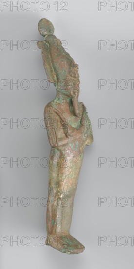 Figurine of Osiris depicted as a mummy-like standing figure wrapped in a shroud. The arms protruding from beneath the shroud are crossed over the chest in the so-called Upper Egyptian configuration. In his right hand, Osiris holds a scepter, while in his left he holds a whip. The head is adorned with an atef crown consisting of the White Crown and two ostrich feathers placed on the sides. The crown is topped with a solar disk and additionally adorned with a large uraeus with two lateral body coils and a long, wavy tail. The shroud fits softly to the body, highlighting the outline of the back and forearms. A false beard is attached to the chin. 664–332 BC; Late Period, Egypt; sculpture; height 17.9 cm, width 5 cm, A small standing figure cast in metal with a worn, textured surface showing a mix of green and brown patina; the figure has a tall headdress with rounded elements, a stylized beard, and arms bent so hands are positioned near the chest; the body appears wrapped or fitted and th...